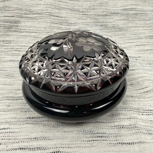 Può includere: Un portagioie rotondo in vetro rosso scuro con coperchio rimovibile. Il coperchio presenta intricati motivi geometrici a stella intagliati in profondità e motivi floreali incisi. La base è liscia, opaca e rosso scuro, in contrasto con la parte superiore sfaccettata.