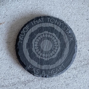 Może przedstawiać: Okrągły, ciemnoszary podkładka z łupka z wygrawerowanym napisem "PROOF THAT TONY STARK HAS A HEART". Podkładka ma koncentryczne wzory okręgów i centralny wzór w kształcie kwiatu. Średnica podkładki wynosi około 10 cm.