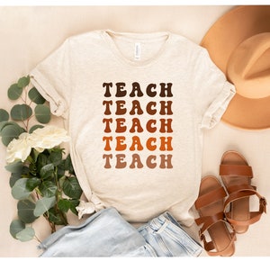 May include: A beige t-shirt with the word "TEACH" repeated five times in a gradient of brown, orange, and red. The shirt is laid flat on a beige surface with a brown hat, blue jeans, and brown sandals.