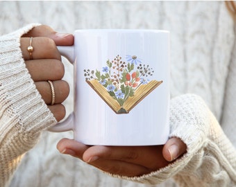 May include: White ceramic mug featuring a watercolor illustration of an open book filled with colorful flowers. The flowers include blue, red, and orange blooms. The mug is held by a person wearing a cream-colored sweater.