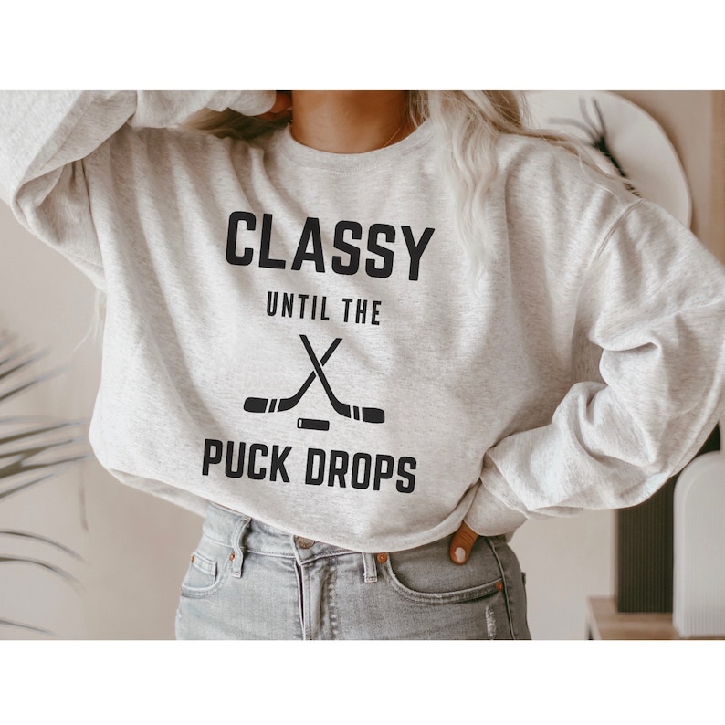 Puede incluir: Una sudadera gris con el texto "Classy Until The Puck Drops" y un gr&aacute;fico de dos palos de hockey cruzados.