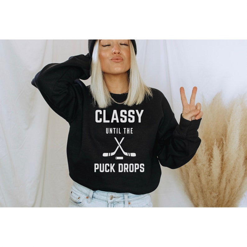 Puede incluir: Sudadera negra con texto blanco que dice "Classy Until The Puck Drops" y un gr&aacute;fico de dos palos de hockey cruzados.