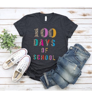 Peut inclure: Un t-shirt gris fonc&eacute; avec le texte "100 DAYS OF SCHOOL" en lettres color&eacute;es avec un motif l&eacute;opard. Les lettres sont soulign&eacute;es de noir.