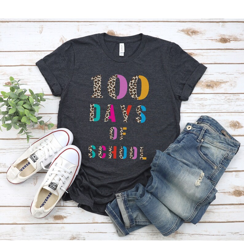 Peut inclure: Un t-shirt gris fonc&eacute; avec le texte "100 DAYS OF SCHOOL" en lettres color&eacute;es avec un motif l&eacute;opard. Les lettres sont soulign&eacute;es de noir.