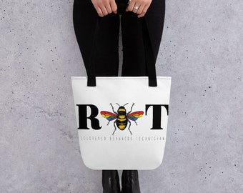 May include: A white tote bag with black handles featuring the large black letters R and T with a bee graphic in the center. The bee has rainbow-colored wings and yellow and black stripes. Below the bee is the text Registered Behavior Technician.