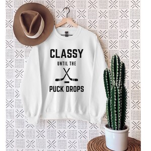 Puede incluir: Sudadera blanca con texto negro que dice "Classy Until the Puck Drops" con un gr&aacute;fico de dos palos de hockey cruzados sobre un disco.