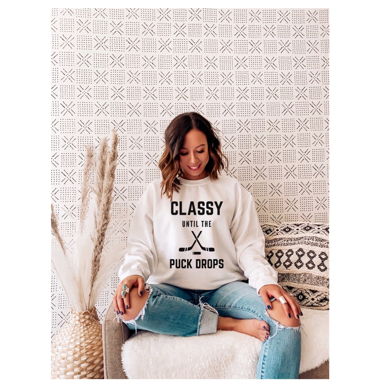 Puede incluir: Sudadera blanca con texto negro que dice "Classy Until the X Puck Drops" y un gr&aacute;fico de dos palos de hockey cruzados.