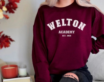Pode incluir: Uma camisa de moletom vinho com texto branco que diz "Welton Academy Est. 1959".