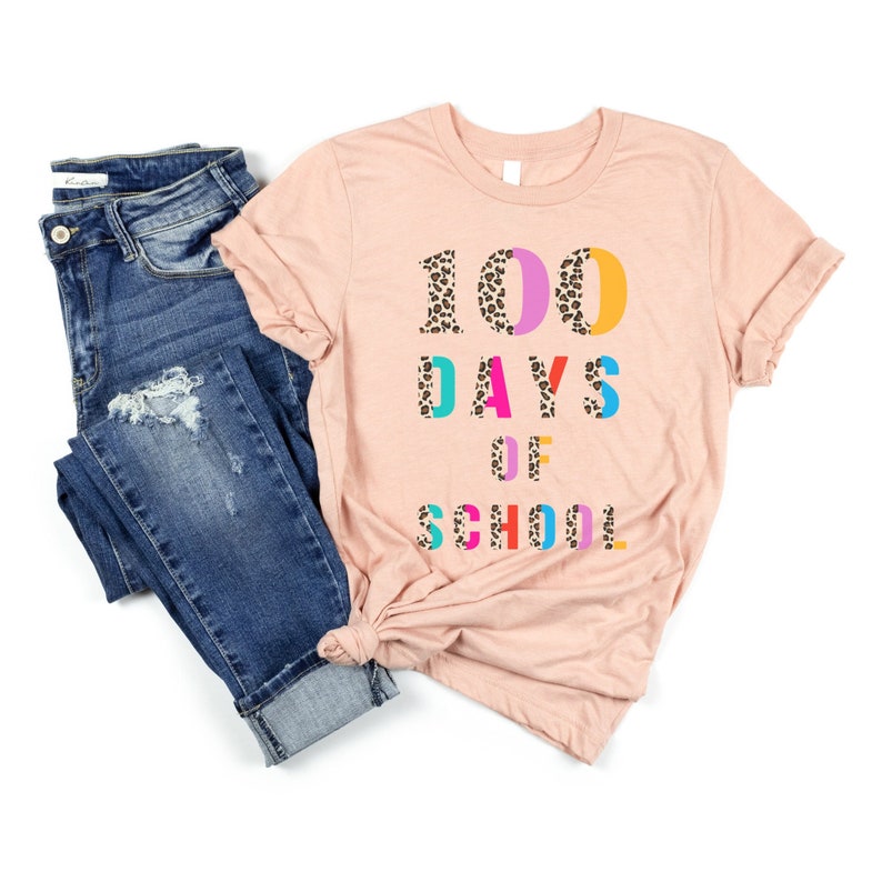 Peut inclure: Un t-shirt rose avec le texte "100 DAYS OF SCHOOL" en lettres color&eacute;es avec un motif l&eacute;opard. Les lettres sont soulign&eacute;es de noir.