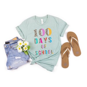 Peut inclure: Un t-shirt bleu clair avec le texte "100 DAYS OF SCHOOL" en lettres color&eacute;es avec un motif l&eacute;opard. Les chiffres 100 sont partiellement recouverts d'un imprim&eacute; l&eacute;opard.