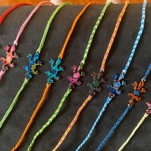 May include: A set of ten colorful braided bracelets with a metal lizard charm on each. The lizards are painted with different patterns and colors, including blue, green, red, orange, yellow, and pink.