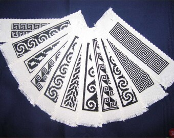 Puede incluir: Un conjunto de marcapáginas de tela blanca con patrones geométricos negros, incluyendo espirales y diseños de llaves. Los marcapáginas están dispuestos en forma de abanico, con un borde deshilachado.