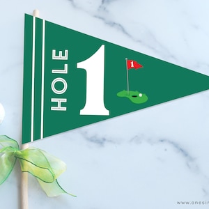 Könnte beinhalten: Ein grünes und weißes Golf-Banner mit dem Text "HOLE 1" und einem grünen Golfplatz mit einer roten Fahne und einem Golfball.