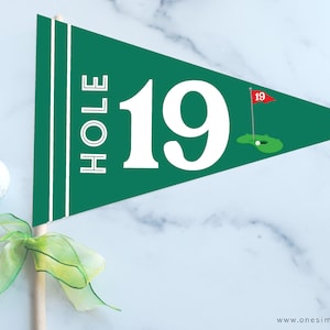 May include: A green pennant flag with the text "HOLE 19" in white, a golf course illustration, and two white stripes. Two golf balls and a green and yellow ribbon are also visible. The flag is on a wooden stick.