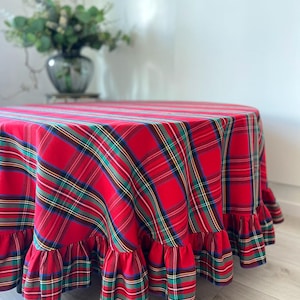 May include: A round tablecloth featuring a vibrant red plaid pattern with intersecting green, yellow, and dark blue stripes. The fabric drapes over a circular surface and is finished with a gathered, ruffled edge that adds texture to the design.