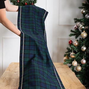 May include: A rectangular table runner featuring a dark green and navy blue plaid pattern. The fabric is bordered by a thin, cream-colored rickrack trim that runs along the length of the runner and across the bottom edge, creating a defined frame.