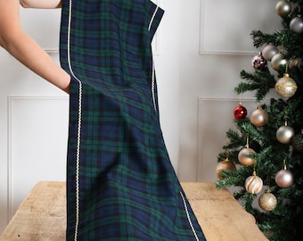 May include: A rectangular table runner featuring a dark green and navy blue plaid pattern. The fabric is bordered by a thin, cream-coloured rickrack trim that runs along the length of the runner and across the bottom edge, creating a defined frame.