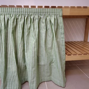 May include: A green and white striped curtain hangs from a wooden shelf. The curtain is made of a light fabric and has vertical stripes. The shelf is made of light-colored wood.