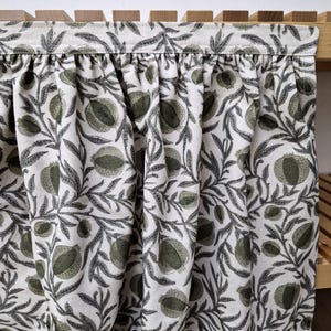 May include: A fabric curtain with a repeating pattern of green fruit and leaves on a cream background. The curtain is gathered at the top and hangs in soft folds. The wooden frame is visible at the top and right side.