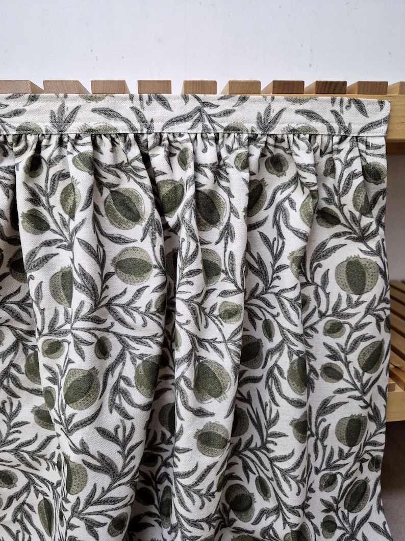 May include: A fabric curtain with a repeating pattern of green fruit and leaves on a cream background. The curtain is gathered at the top and hangs in soft folds. The wooden frame is visible at the top and right side.