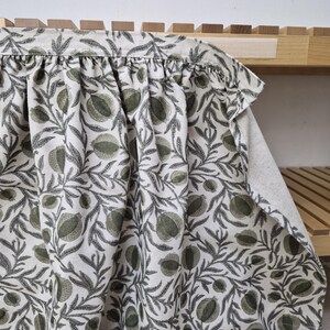 May include: A white fabric with a repeating pattern of green pomegranates and gray leaves. The fabric is draped over a wooden shelf, showcasing the design and texture. The fabric's edge is gathered, adding a decorative element.