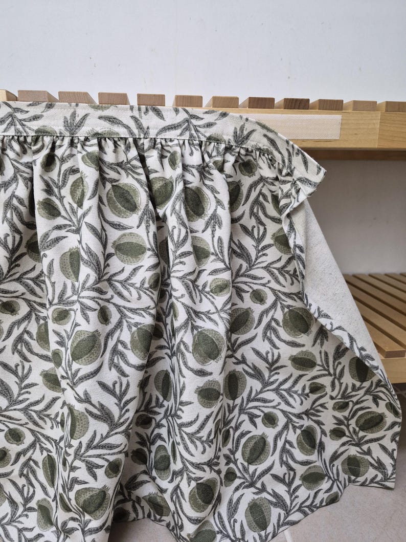 May include: A white fabric with a repeating pattern of green pomegranates and gray leaves. The fabric is draped over a wooden shelf, showcasing the design and texture. The fabric's edge is gathered, adding a decorative element.