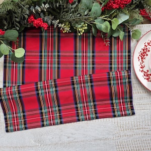 May include: A rectangular fabric table runner featuring a red, green, blue, and white plaid pattern. The runner is folded over itself, displaying the consistent tartan design. A matching folded napkin sits on a white plate nearby.