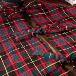 May include: Close-up of a red, green, black, and yellow plaid Christmas tree skirt. The skirt is made of fabric and has ties to secure it around the tree base. The plaid pattern is a classic holiday design.