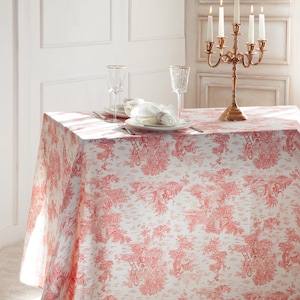 Puede incluir: Un mantel blanco con un patrón de toile rojo que presenta un diseño repetitivo de árboles, pájaros y casas. Dos copas de vino de vidrio transparente y un candelabro dorado con velas encendidas están sobre la mesa.