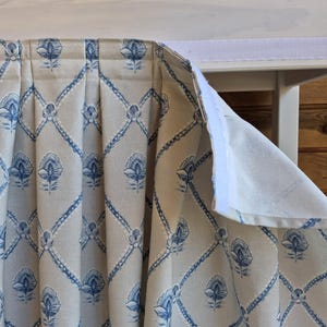 May include: Cream-coloured fabric with a blue floral and diamond pattern. The fabric is pleated and attached to a white surface with a velcro strip. The design features stylised flowers and a trellis-like grid.