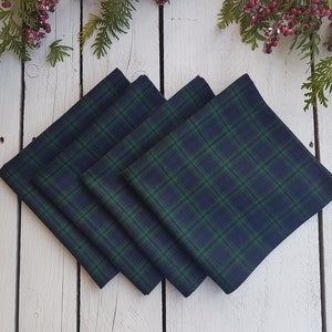 Può includere: Quattro tovaglioli quadrati di stoffa con un motivo a quadri blu navy e verde. I tovaglioli sono impilati in diagonale su una superficie di legno bianco, con vegetazione e bacche rosse sullo sfondo.