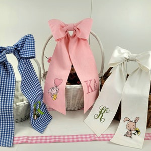 Könnte beinhalten: Drei weiße Weidenkörbe mit verschiedenen Stoffschleifen und personalisierter Stickerei. Die Schleifen sind blau-weiß kariert, rosa und weiß. Die Stickerei umfasst ein Häschen mit einem Ballon, ein Häschen mit einer Blume und die Initialen "K.A.M."
