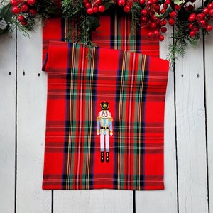 May include: Red and green plaid table runner with an embroidered nutcracker design. The nutcracker is blue and white with a gold hat.
