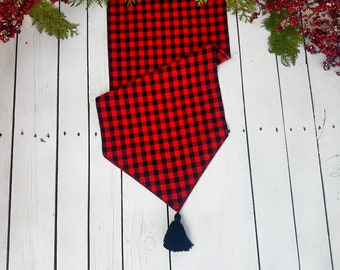 May include: Red and black checkered table runner with a black tassel. The runner is on a white wooden surface.