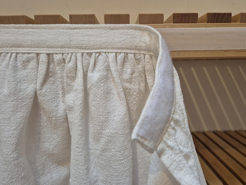 May include: White cotton fabric with a ruffled edge and a hook and loop fastener. The fabric is hanging from a wooden shelf.