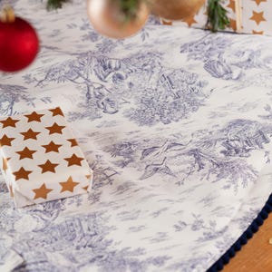 May include: A white Christmas tree skirt with a blue toile pattern depicting pastoral scenes. The skirt has a dark blue pom-pom trim and a wrapped gift with gold star accents. Ornaments hang above.