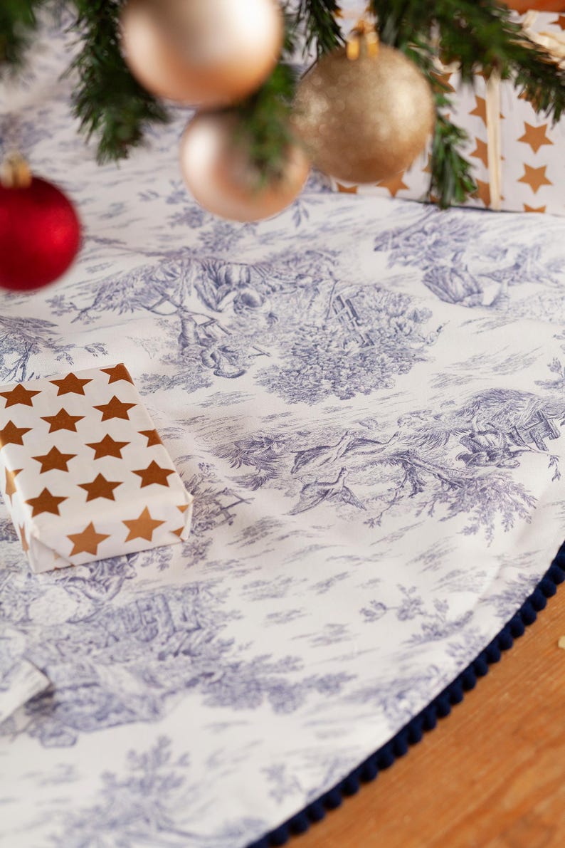 May include: A white Christmas tree skirt with a blue toile pattern depicting pastoral scenes. The skirt has a dark blue pom-pom trim and a wrapped gift with gold star accents. Ornaments hang above.