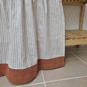 May include: A beige and white striped curtain with a rust-colored border. The curtain is hanging in front of a wooden shelf. The fabric has a textured appearance and is slightly gathered at the top.