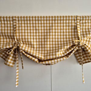 Könnte beinhalten: Senfgelbe und weiße Vichy-Karo-Gardinenblende mit Bindebändern. Der Stoff ist über eine Holzstange drapiert und erzeugt einen weichen, gerafften Effekt. Diese Fensterdekoration verleiht jedem Raum einen Hauch von Landhausstil.