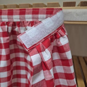 Op de afbeelding: Rood en wit gingham-stof met een witte klittenbandsluiting. De stof is aan de bovenkant geplooid en heeft een gerimpelde rand.