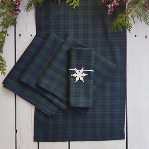 May include: A table runner and matching napkins in a green and navy blue plaid pattern. A decorative snowflake napkin ring is on one of the napkins. The items are arranged on a white wooden surface, with greenery and berries in the background.