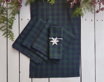 May include: A set of green and navy blue plaid table runner and napkins. A snowflake napkin ring adorns one napkin. The items are arranged on a white wooden surface, with sprigs of greenery and berries in the background, creating a festive look.
