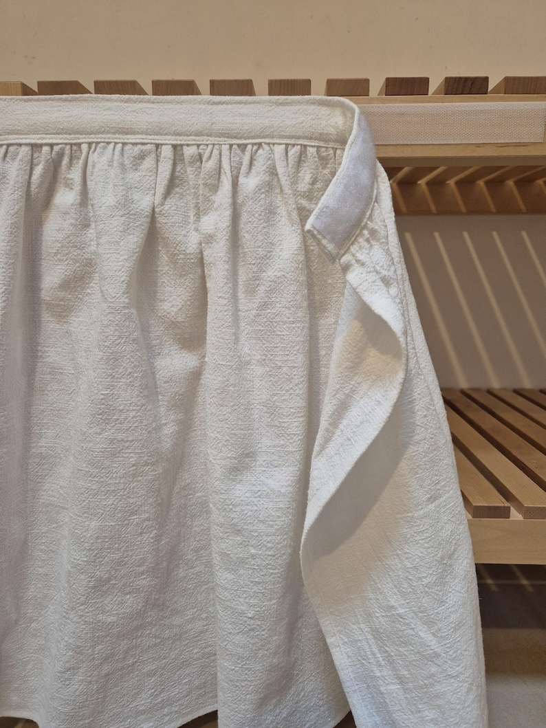 May include: A white long-sleeved shirt with a gathered neckline, hanging on a wooden shelf.