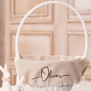 May include: White wicker basket with a linen liner and a white handle. The liner has the name "Oliver" printed on it. Two white ceramic bunny figurines are in front of the basket.
