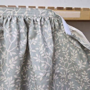 May include: A pale blue fabric featuring a white leaf design, gathered at the top. The fabric is attached to a wooden frame with a white Velcro fastening. The pattern consists of repeating leaves and branches.