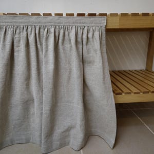 May include: A light gray linen fabric skirt, gathered at the top, hangs in front of a wooden shelf. The skirt has a simple, natural aesthetic, with a slightly textured appearance. The wooden shelf has a natural finish.