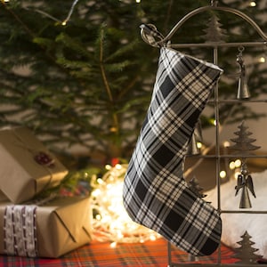 May include: A black and white plaid Christmas stocking hangs from a decorative metal stand with small bells and tree ornaments. Presents wrapped in brown paper with ribbons and a Christmas tree are in the background.