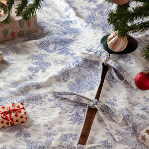 May include: A white and blue toile Christmas tree skirt with a detailed pastoral scene design. The skirt has a dark blue trim and ties to secure it around the tree. Ornaments and wrapped gifts are visible.