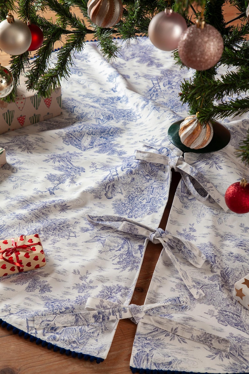 May include: A white and blue toile Christmas tree skirt with a detailed pastoral scene design. The skirt has a dark blue trim and ties to secure it around the tree. Ornaments and wrapped gifts are visible.
