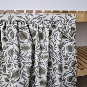 May include: A close-up of a fabric curtain with a botanical pattern. The curtain features a white background with a repeating design of green pomegranates and gray leaves. The curtain is attached to a wooden shelf.